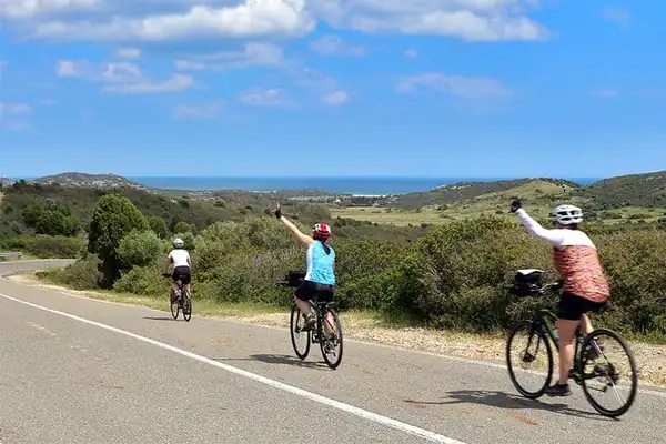 Two cyclists waving on a coastal road in Sardinia